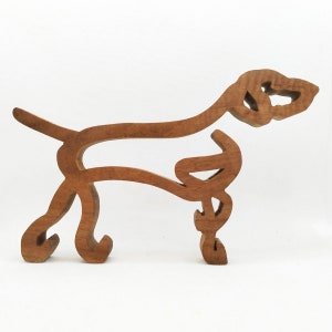 Pode incluir: Um recorte de madeira de um cão em um design estilizado e abstrato. O cão está em pé sobre suas quatro patas com a cabeça virada para o lado. O recorte é feito de madeira marrom claro.