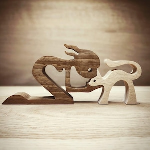 Könnte beinhalten: Holzskulptur, die eine stilisierte Figur darstellt, die mit einem kleineren Tier, möglicherweise einer Katze, interagiert. Die größere Figur ist dunkelbraun, während die kleinere hellbeige ist. Die Figuren sind vor einem hellbraunen Hintergrund platziert.