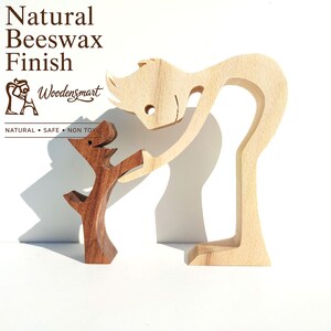 Può includere: Due statuette in legno stilizzate con finitura in cera d'api naturale. Una figura più grande di colore chiaro si sporge verso una figura più piccola di colore scuro. Il testo recita: Natural Beeswax Finish, Woodensmart, NATURAL • SAFE • NON TOXIC.