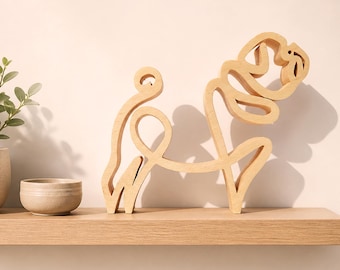 Op de afbeelding: Een lichtgekleurde houten sculptuur van een hond in minimalistische stijl. De hond is afgebeeld in een doorlopende lijn, met een gekrulde staart en kop. De sculptuur staat op een houten plank, met een kleine keramische kom en een plant op de achtergrond.
