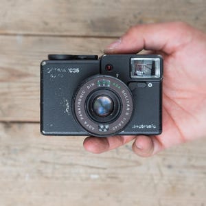 May include: A black Optima C35 camera with a Solitar lens, held in a hand. The camera has a flash and various dials. The lens has the text "AGFA PARATRONIC DIN ASA SOLITAR S 1:2.8/40". The camera is on a wooden surface.