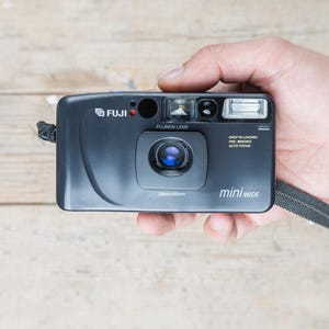 Op de afbeelding: Een zwarte Fuji Mini Wide filmcamera met ingebouwde flitser en polsband. De camera heeft een lens met de tekst "FUJINON LENS" en "28mm/45mm" erop. De camera wordt in een hand gehouden.