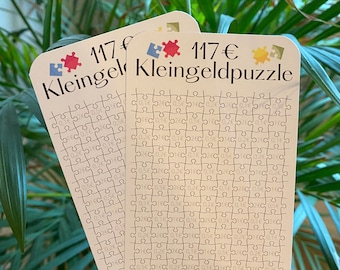 May include: Two white cards with a puzzle design. The cards have the text "117 € Kleingeldpuzzle" printed on them. The puzzle design is made up of small squares with a euro symbol in each square.