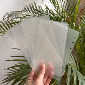 以下が含まれることがあります： タブと穴の開いた半透明のプラスチック製バインダーディバイダーのスタック。ディバイダーは、緑の葉の植物の背景の前に保持されています。ディバイダーは、ドキュメントを整理するように設計されています。