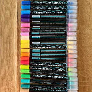 Può includere: Una collezione di pennarelli per pittura acrilica in vari colori, tra cui nero, bianco, blu, rosa, giallo, arancione, rosso e verde. Ogni pennarello ha un corpo nero e un cappuccio trasparente. Il testo "ACRYLIC PAINT MARKER" è visibile.