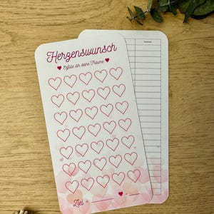 Può includere: Un tracker di cuori stampabile rosa e bianco con il testo "Herzenswunsch" e "Erfülle dir deine Träume" in rosa. Il tracker ha 48 cuori da colorare e uno spazio per scrivere un obiettivo.