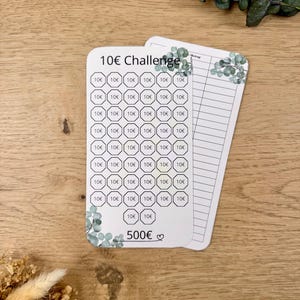 Könnte beinhalten: Eine weiße Karte mit einer 10€-Herausforderung darauf gedruckt. Die Karte hat ein Raster mit 50 Kreisen, in denen jeweils 10€ gedruckt sind. Die Karte hat auch ein florales Design mit Eukalyptusblättern. Die Karte hat insgesamt 500€ darauf gedruckt.