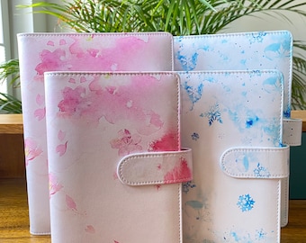 May include: Four different patterned white binders with a flap closure. The binders have watercolour designs of pink flowers, blue snowflakes, and blue and white abstract patterns.