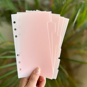 May include: A stack of pink and white binder dividers with tabbed edges and three-hole punches. The dividers have a gradient color effect, transitioning from white to pink. The dividers are held in a hand against a blurred green background.