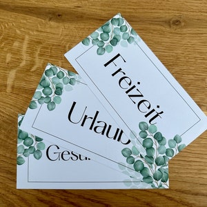 Può includere: Biglietti rettangolari bianchi con testo nero e disegni ad angolo con foglie di eucalipto verdi. I biglietti riportano le parole "Freizeit", "Urlaub" e "Gesund". I biglietti sono disposti su una superficie di legno.