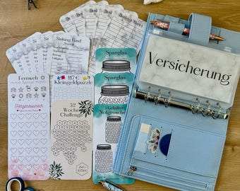 Könnte beinhalten: Ein hellblauer Ordner mit verschiedenen Finanzplanungswerkzeugen, darunter Budgetblätter, Spar-Tracker und Aufkleber. Das Bild zeigt auch Stifte, Washi-Tape und einen Regenbogen-Schlüsselanhänger, alles auf einer Holzoberfläche angeordnet.