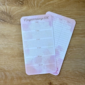 Op de afbeelding: Twee roze en witte spaaruitdagingskaarten. Eén kaart heeft de tekst "Minigeldchallenge 83€" en een raster om de besparingen bij te houden. De andere kaart heeft een aquarelontwerp en lijnen om informatie vast te leggen.