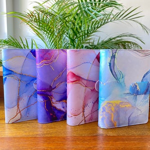 Pode incluir: Quatro cadernos de couro coloridos com designs abstratos de aquarela e detalhes dourados. Os cadernos têm uma capa azul, roxa, rosa e azul claro.