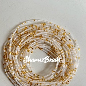 May include: White and gold beaded waist beads. The beads are small and round, and they are strung together in a long strand. The beads are arranged in a pattern of white and gold.