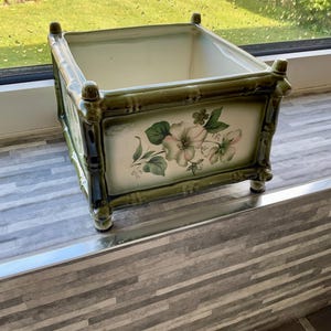 May include: A square green and white ceramic planter with a bamboo design and floral pattern. The planter has a white interior and is sitting on a tiled surface.