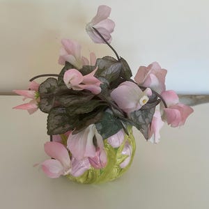 May include: A small, round, lime green ceramic vase with a raised, sculpted leaf pattern. The vase is filled with a cluster of delicate, light pink cyclamen flowers featuring dark green, heart-shaped leaves with silver-grey variegated patterns.