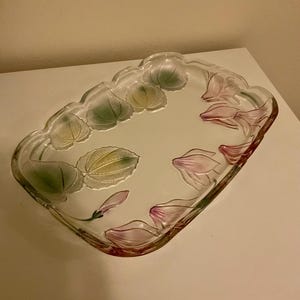 May include: A clear, rectangular glass serving tray with a scalloped edge. The tray is decorated with painted pink and green floral designs. The flowers and leaves are arranged around the edges of the tray.