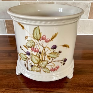 May include: A white ceramic utensil holder with a floral design featuring green leaves, pink berries, and blackberries. The holder has three small feet and a scalloped rim.