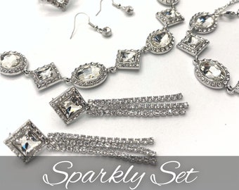 May include: A sparkly jewelry set with a necklace, earrings, and a ring. The set features clear crystal stones in various shapes, including round, oval, and square. The necklace and earrings have a silver tone finish.