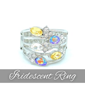 May include: A silver ring with a multi-layered design featuring iridescent and yellow stones. The ring is adorned with clear rhinestones.