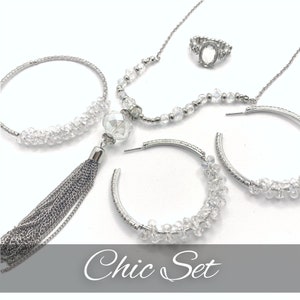 May include: A jewelry set featuring a silver-tone necklace with a large faceted crystal pendant and a long chain tassel, a pair of silver hoop earrings with clear crystal beads, a beaded bracelet, and a ring with an oval setting. Text reads Chic Set.