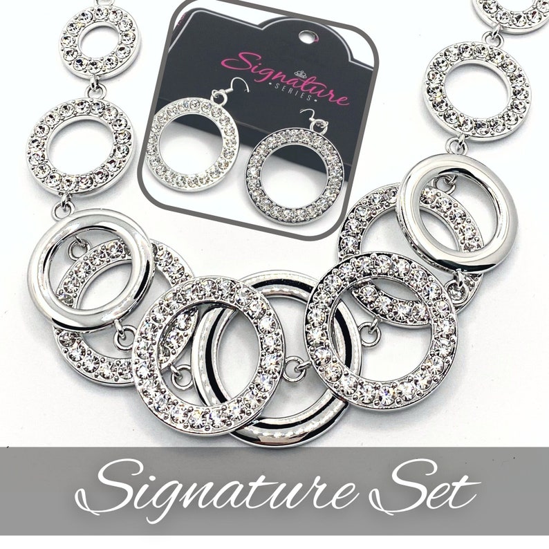 May include: A silver-toned jewelry set featuring a necklace and matching earrings. The necklace and earrings have a circular design, with the circles adorned with sparkling crystals. The earrings are dangling. The set is displayed on a white background. The text "Signature Set" is visible.