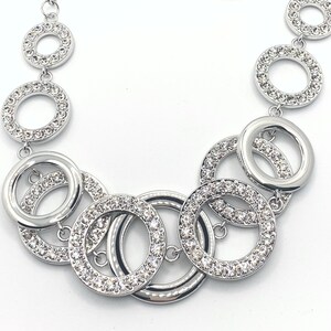 May include: Silver-toned necklace featuring a series of interconnected open circles. Some circles are encrusted with small, sparkling crystals, adding a touch of glamour. The necklace has a polished finish and a delicate chain.