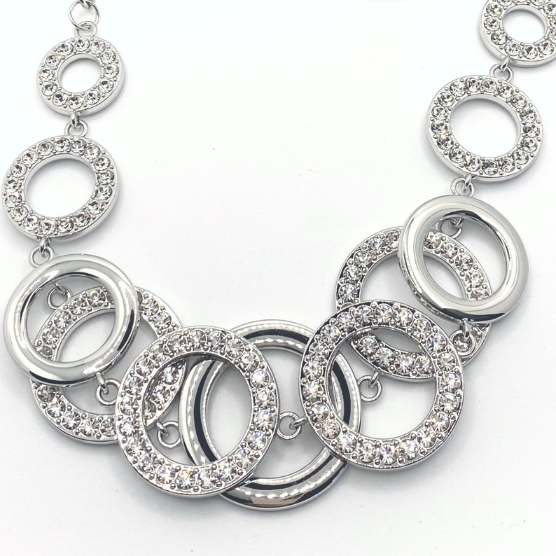 May include: Silver-toned necklace featuring a series of interconnected open circles. Some circles are encrusted with small, sparkling crystals, adding a touch of glamour. The necklace has a polished finish and a delicate chain.