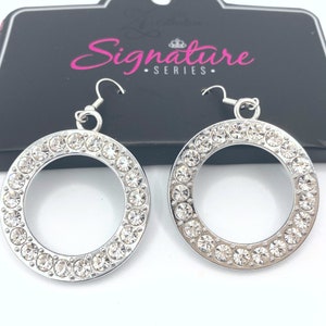 May include: A pair of silver-toned hoop earrings. Each earring features a circle of clear, sparkling rhinestones set around the outer edge. The earrings have a simple hook closure. The packaging behind the earrings reads "Signature Series."
