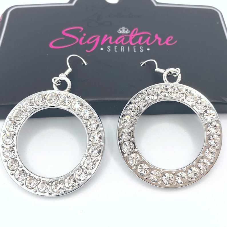 May include: A pair of silver-toned hoop earrings. Each earring features a circle of clear, sparkling rhinestones set around the outer edge. The earrings have a simple hook closure. The packaging behind the earrings reads "Signature Series."