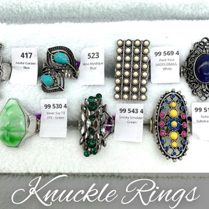 May include: A collection of silver knuckle rings with various colored stones and designs. The rings include turquoise, green, white, blue, and yellow stones. The text on the labels reads: "Adobe Garden Blue", "Mesa Mystique Blue", "Pack Your SADDLEBAGS-White", "Once Upon Meadow-E", "Never Say TIE DYE-Green", "Smoky Smolder - Green", "Sonoran Solstice - Blue".