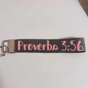Può includere: Un portachiavi grigio con la scritta "Proverbi 3:56" in rosa. Il cinturino ha una chiusura e un portachiavi in metallo color argento.