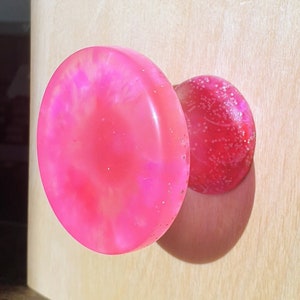 May include: A pink and white resin cabinet knob with a round shape and a slightly domed top.