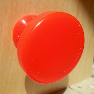 May include: A bright red plastic knob with a smooth, round surface. The knob has a small, raised lip around the edge.