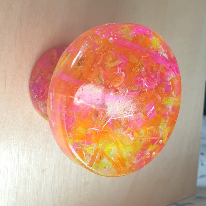 May include: A round, clear resin coaster with embedded pink, yellow, and orange glitter. The coaster has a slightly raised edge.