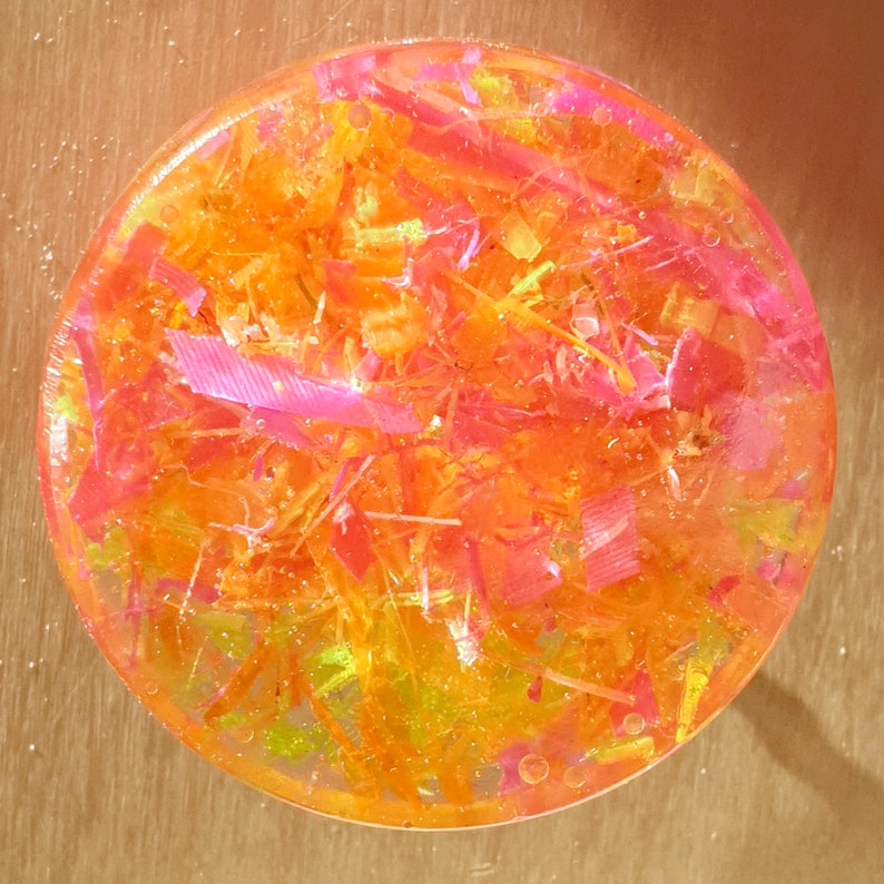 May include: A round, clear resin coaster with embedded pink, orange, and yellow glitter. The glitter is arranged in a random pattern and creates a sparkly effect.