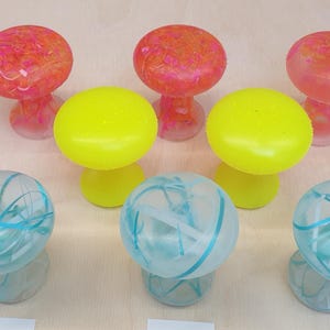 Könnte beinhalten: Eine Sammlung von bunten, pilzförmigen Dekorationsobjekten aus Harz. Die Pilze sind in Gelb-, Rosa- und Blautönen mit durchgehenden Farbwirbeln.