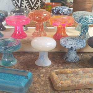Peut inclure: Une collection de 14 boutons en résine en forme de champignon de différentes couleurs, motifs et textures. Les boutons sont disposés sur une surface brune avec deux petits plateaux rectangulaires.