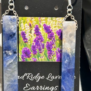 Peut inclure: Boucles d'oreilles avec des pendentifs rectangulaires en pierre bleue et blanche, reli&eacute;s par des cha&icirc;nes et des crochets argent&eacute;s. Le fond pr&eacute;sente une carte avec une image de champ de lavande et le texte "Red Ridge Lavender Earrings".