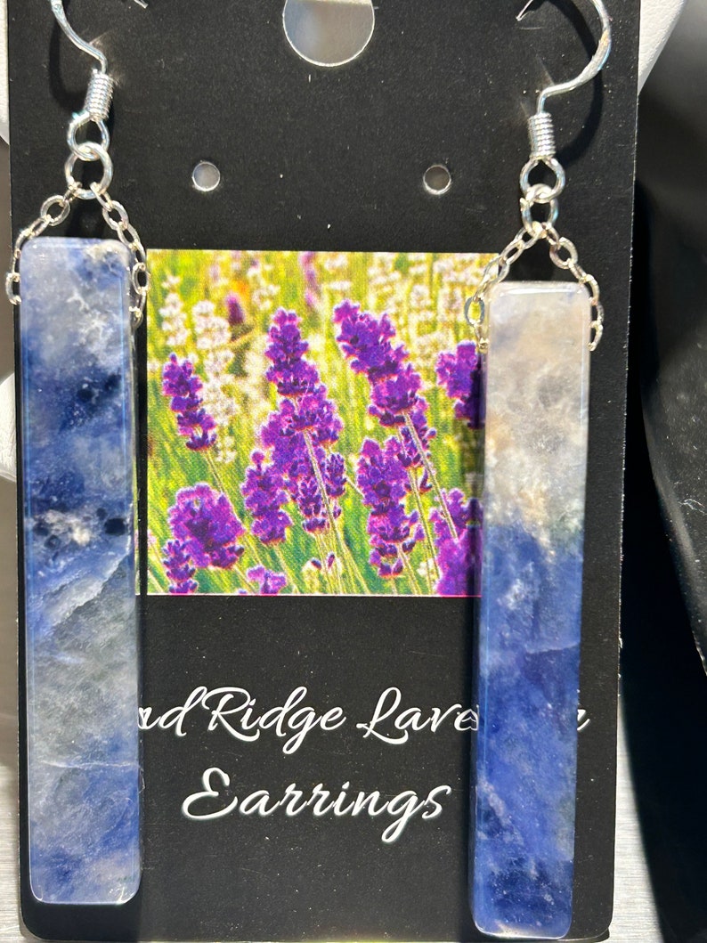 Peut inclure: Boucles d'oreilles avec des pendentifs rectangulaires en pierre bleue et blanche, reli&eacute;s par des cha&icirc;nes et des crochets argent&eacute;s. Le fond pr&eacute;sente une carte avec une image de champ de lavande et le texte "Red Ridge Lavender Earrings".