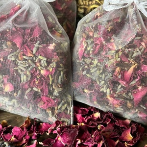 May include: Two white mesh bags filled with dried rose petals and lavender. The bags are on a brown surface with loose rose petals scattered around them.