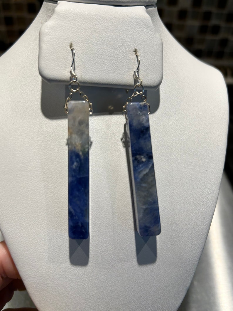 Peut inclure: Une paire de boucles d'oreilles rectangulaires avec un motif marbr&eacute; bleu et blanc. Les boucles d'oreilles sont suspendues &agrave; des crochets et des cha&icirc;nes de couleur argent&eacute;e. Les boucles d'oreilles sont expos&eacute;es sur un pr&eacute;sentoir &agrave; bijoux blanc.