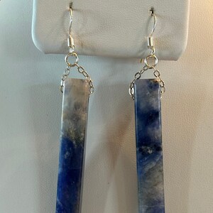 Peut inclure: Une paire de boucles d'oreilles rectangulaires avec un d&eacute;grad&eacute; de tons bleus et gris. Les boucles d'oreilles sont suspendues &agrave; des crochets et des cha&icirc;nes de couleur argent&eacute;e. Les boucles d'oreilles sont expos&eacute;es sur un pr&eacute;sentoir &agrave; bijoux blanc.