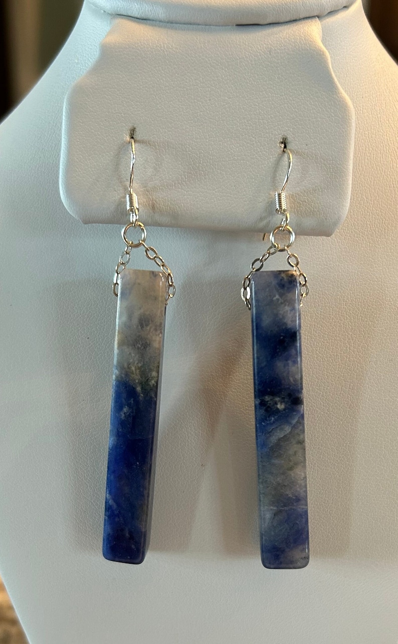 Peut inclure: Une paire de boucles d'oreilles rectangulaires avec un d&eacute;grad&eacute; de tons bleus et gris. Les boucles d'oreilles sont suspendues &agrave; des crochets et des cha&icirc;nes de couleur argent&eacute;e. Les boucles d'oreilles sont expos&eacute;es sur un pr&eacute;sentoir &agrave; bijoux blanc.