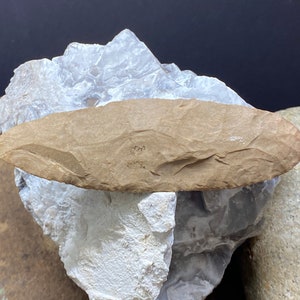 May include: A light brown, oval-shaped, stone tool with a smooth, slightly textured surface. The tool is resting on a white, rocky surface.