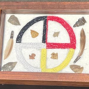 May include: A framed artwork featuring a beaded Native American medicine wheel with four colors: black, white, red, and yellow. The wheel is surrounded by feathers and other natural elements.