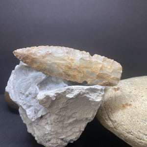 May include: A light brown and white arrowhead made of stone, resting on a white rock. The arrowhead is pointed and has a rough texture.