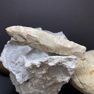 May include: A light brown, flint arrowhead with a pointed tip and a rough, textured surface. The arrowhead is resting on a pile of white rocks.