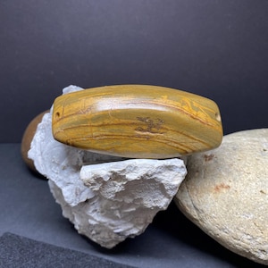 Puede incluir: Una cuenta de piedra lisa de forma ovalada con un patrón de mármol marrón y amarillo. La cuenta tiene un agujero perforado, lo que sugiere que se usó para joyería u otros fines decorativos.