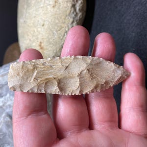 Puede incluir: Una punta de flecha de color marrón claro con una superficie rugosa y texturizada. La punta de flecha se sostiene en una mano.